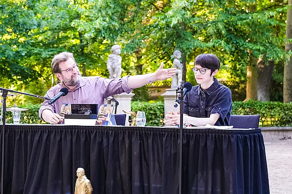 Zwei Personen sitzen an einem Tisch im Freien. Eine Person spricht und gestikuliert, während die andere aufmerksam zuhört. Im Hintergrund sind Bäume und Statuen sichtbar. Auf dem Tisch liegen Bücher und Gläser.