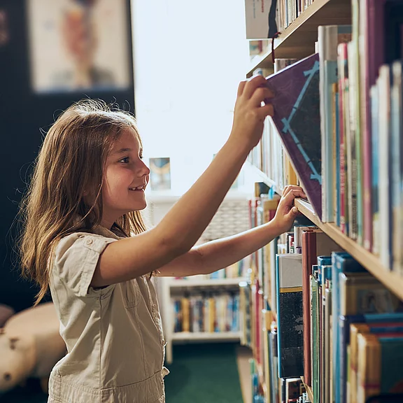 Ein Mädchen steht an einem Bücherregal in einer Bibliothek. Sie lächelt und greift nach einem Buch auf dem Regal. Im Hintergrund sind weitere Bücher und ein heller Raum zu sehen.