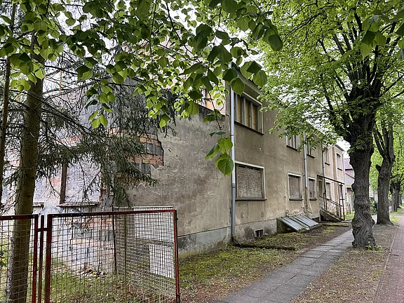 Verlassener, heruntergekommener Wohnblock an einer baumbestandenen Straße. Fenster sind mit Brettern vernagelt, Fassade rissig und verwittert. Ein schmaler Gehweg führt am Gebäude vorbei, im Vordergrund ein einfaches Metalltor. Szene wirkt still und vernachlässigt.