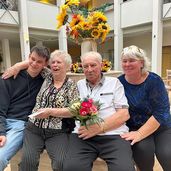 Vier Menschen sitzen eng beieinander und lachen. Eine ältere Frau umarmt einen jungen Mann. Daneben hält ein älterer Mann einen bunten Blumenstrauß, eine weitere Frau sitzt lächelnd neben ihm. Feierliche, freundliche Stimmung in einem hellen Raum mit Blumen.