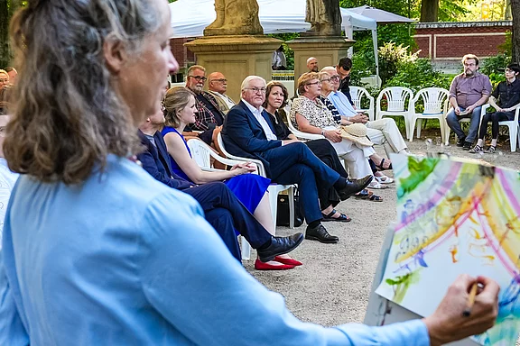 Eine Künstlerin malt an einer Leinwand, während eine Gruppe von Menschen sitzt und zuschaut. Die Zuschauer sind in einem parkähnlichen Ambiente mit Stühlen platziert. Die Szene vermittelt eine kreative und gemeinschaftliche Atmosphäre.