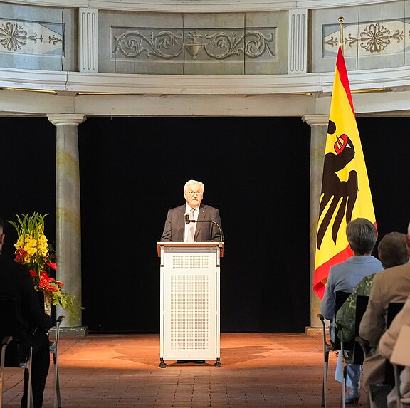 Ein älterer Mann steht hinter einem Rednerpult und hält eine Rede. Im Hintergrund sind zwei Fahnen zu sehen, eine davon mit dem Wappen eines Bären. Vor ihm sitzen Zuhörer, die ihm aufmerksam lauschen. Die Szenerie wirkt feierlich und respektvoll.