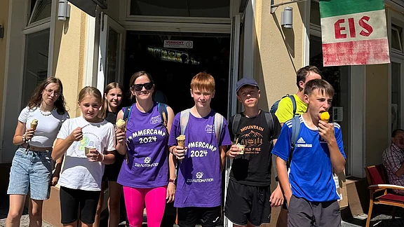 Eine Gruppe Jugendlicher steht vor einer Eisdiele und isst Eis. Sie lachen, halten Becher und Waffeln, sommerliche Kleidung. Über dem Eingang steht „Il Gelato“, daneben ein „EIS“-Schild. Sonniges Wetter, entspannte Stimmung.