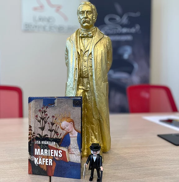 Auf einem Tisch: goldene Männerstatue. Davor das Buch „MARIENSKÄFER“ von Lisa Kränzler mit mittelalterlichem Motiv auf dem Cover. Daneben eine kleine Spielfigur im schwarzen Anzug mit Zylinder.