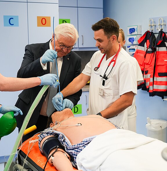 Der deutsche Bundespräsident steht in einem medizinischen Raum neben einem Arzt, der eine Notfallbehandlung an einem Patienten durchführt. Beide tragen Handschuhe und sind konzentriert bei der Arbeit, während ein weiteres Präsenzpersonal im Hintergrund zu sehen ist.