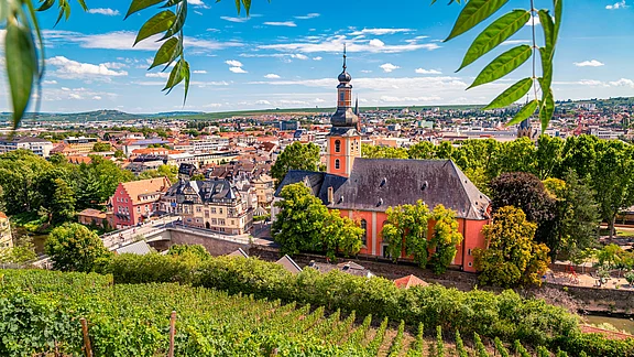 Ein malerischer Blick auf die Stadt, umgeben von Weinbergen. Im Vordergrund ist eine große, orangefarbene Kirche mit einer hohen Spitze, während sich dahinter eine Vielzahl von Gebäuden und grünen Bäumen erstreckt. Der Himmel ist blau mit einigen Wolken.