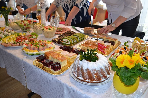 Reich gedecktes Buffet: verschiedene Kuchen und Torten, Stückkuchen mit Gelee, Biskuitrolle, belegte Häppchen und Salate. Ein Puderzucker-Gugelhupf vorne. Hände schneiden und teilen Speisen. Festliche, gesellige Atmosphäre.