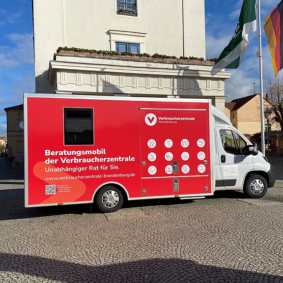 Rotes Beratungsmobil der Verbraucherzentrale Brandenburg auf einem Platz. Text am Fahrzeug: „Beratungsmobil der Verbraucherzentrale – Unabhängiger Rat für Sie“. Sichtbar sind Symbole zu Beratungsthemen und eine Webadresse. Kernaussage: mobile Verbraucherberatung vor Ort.