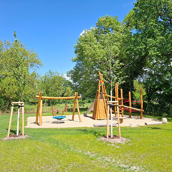 Ein Spielplatz im Freien mit einer Holzschaukel, einer Nestschaukel und einem Spielturm, umgeben von grünen Wiesen und Bäumen. Der Himmel ist blau und die Sonne scheint, was eine einladende Atmosphäre schafft.