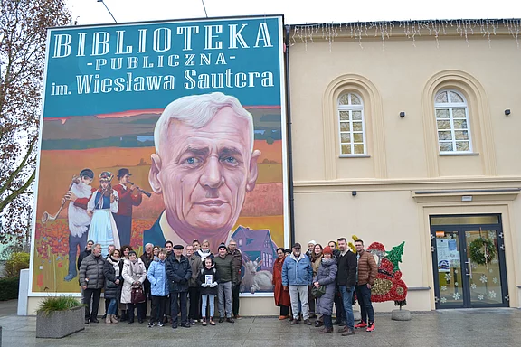 Gruppe von Menschen posiert vor einer Bibliothek mit riesigem Wandgemälde: ein Porträt eines älteren Mannes, daneben ländliche Szene. Auf der Fassade steht groß „Biblioteka Publiczna“. Gruppenfoto vor dem Gebäude.