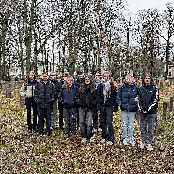 Eine Gruppe von 13 Jugendlichen posiert in einem Friedhof mit Bäumen im Hintergrund. Sie tragen warme Winterkleidung und stehen in einer lockeren Formation auf einem mit Laub bedeckten Boden. Der Ort wirkt ruhig und nachdenklich.