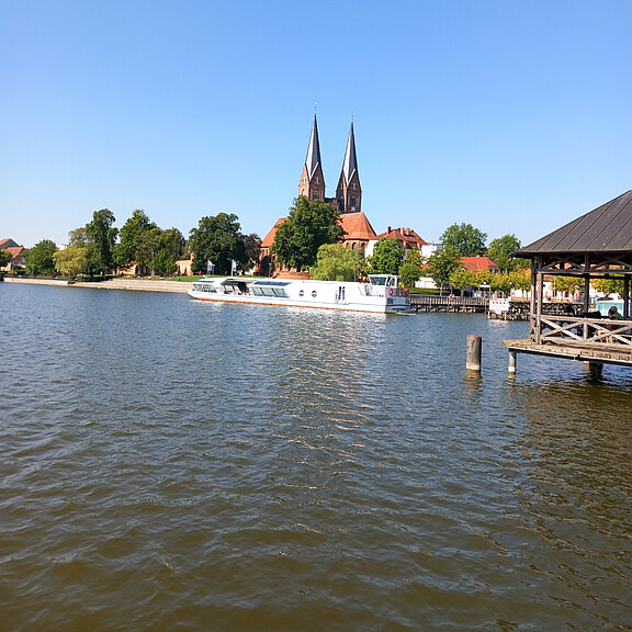 Ein ruhiger Fluss mit einem Ausflugsschiff im Vordergrund. Im Hintergrund sind grüne Bäume und ein markantes Gebäude mit zwei hohen Türmen zu sehen. Der Himmel ist klar und blau, was eine friedliche Atmosphäre schafft.