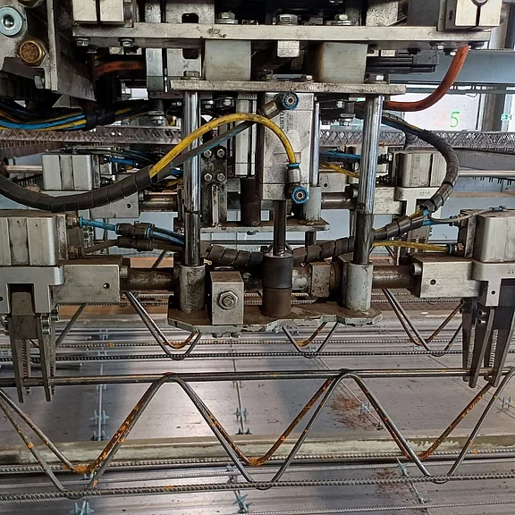 Nahaufnahme einer automatisierten Punktschweißmaschine: Mechanische Führungen, Pneumatikschläuche und Elektroden arbeiten über zickzackförmigen Stahldrähten, um ein Bewehrungsgitter zu verbinden.