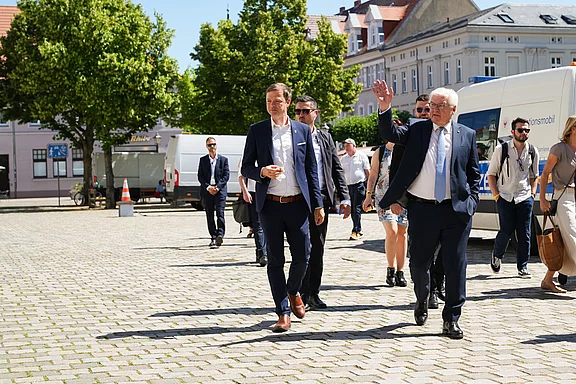 Eine Gruppe von Menschen geht auf einem Pflasterweg. Einige tragen Anzüge, andere lässige Kleidung. Ein Mann mit gestrecktem Arm winkt in Richtung der Kamera. Umgeben von Bäumen und Gebäuden wirkt die Szene freundlich und einladend.