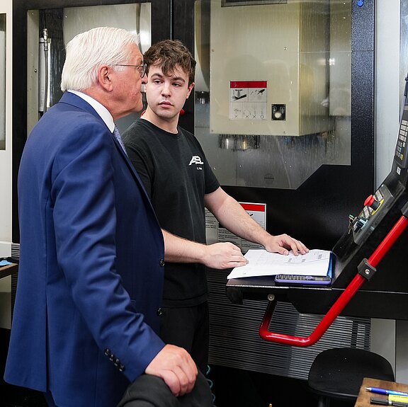 Ein älterer Mann in einem Anzug spricht mit einem jüngeren Mann, der Arbeitskleidung trägt. Sie stehen in einer Werkstatt vor einer Maschine und schauen auf ein Dokument, das auf einem Tisch liegt. Die Szene zeigt den Austausch von Wissen und Erfahrung in einem industriellen Umfeld.