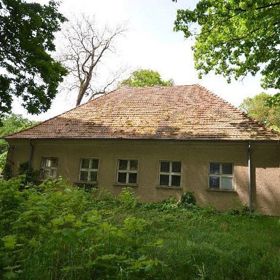 Ein kleines, einstöckiges Landhaus mit steilem Ziegeldach und mehreren kleinen Fenstern, verputzte Fassade. Von dichtem, verwildertem Grün und Bäumen umgeben; wirkt alt und ländlich.