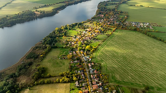 Luftaufnahme eines ländlichen Gebiets mit einem Fluss, der durch die Landschaft fließt. Auf der einen Seite sind Wiesen und Felder, auf der anderen Seite eine kleine Ansiedlung mit Wohnhäusern und Bäumen. Die Natur zeigt herbstliche Farben.
