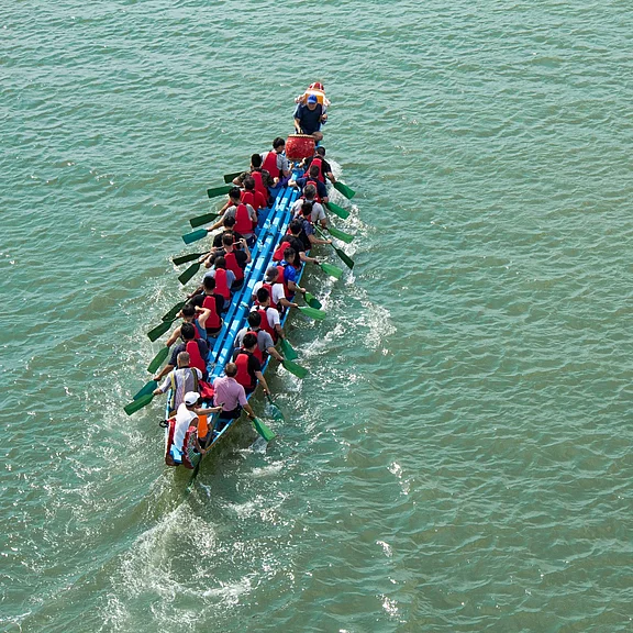 Luftaufnahme eines langen Drachenbootes auf ruhigem Wasser. Zahlreiche Paddler in Reihen rudern synchron nach vorne, ein Steuermann steht am Heck. Spritzwasser hinter dem Boot zeigt die Bewegung.