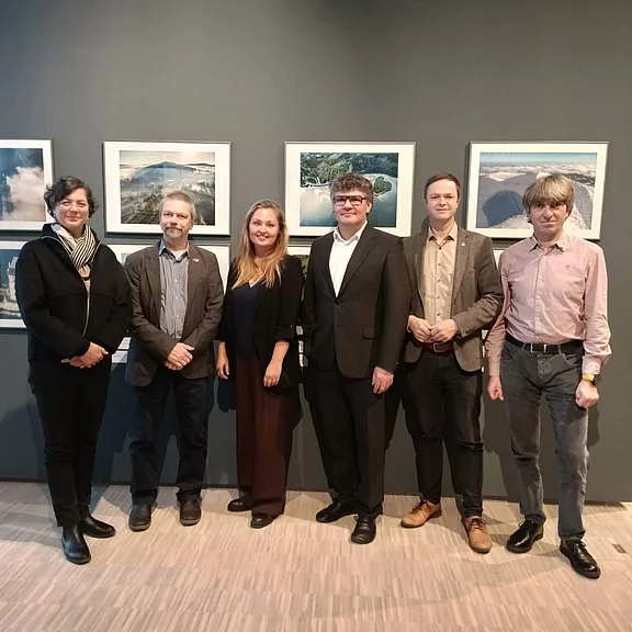Sechs Erwachsene stehen nebeneinander in einer Galerie vor gerahmten Landschaftsfotos. Sie blicken zur Kamera und posieren für ein Gruppenfoto, überwiegend in formeller Kleidung.
