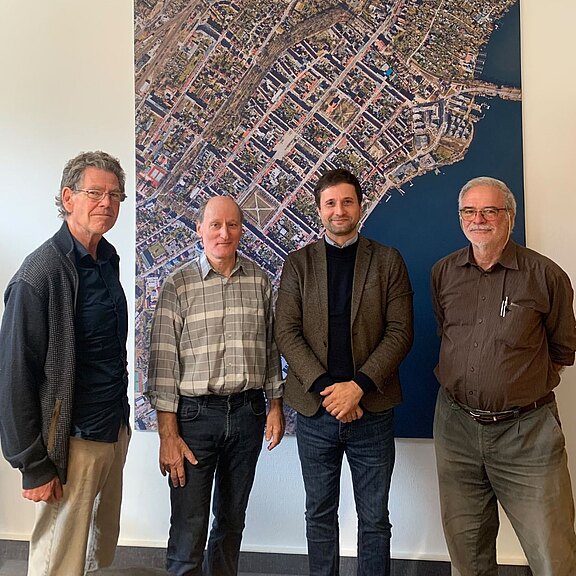 Gruppenfoto von vier Männern in einem Büro vor einer großformatigen Luftaufnahme einer Stadt am Wasser; alle blicken in die Kamera.