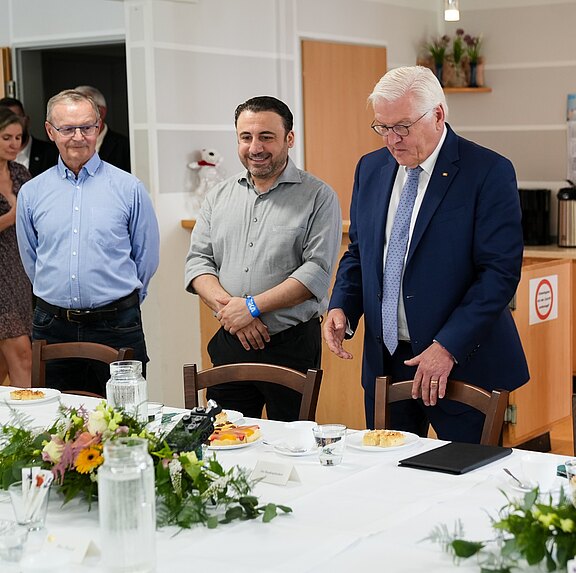 Drei Männer stehen um einen gedeckten Tisch, der mit Blumen und Speisen dekoriert ist. Zwei Männer tragen formelle Kleidung, während der dritte leger gekleidet ist. Alle blicken auf den Tisch, es herrscht eine freundliche Atmosphäre.