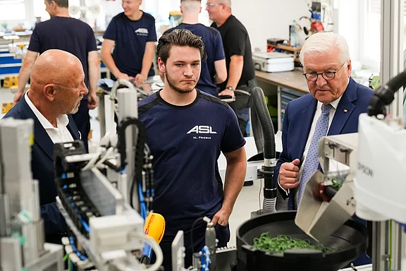 Der Bundespräsident steht in einer Werkstatt im Gespräch mit zwei Männern, während sie eine Maschine betrachten. Einer der Männer trägt ein T-Shirt mit der Aufschrift "ASL". Im Hintergrund sind weitere Personen und Maschinen zu sehen, die für die Produktion verwendet werden.