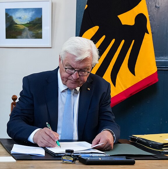 Ein älterer Mann mit Brille, bekleidet mit einem dunklen Anzug und hellblauer Krawatte, sitzt an einem Tisch und schreibt auf einem Blatt Papier. Im Hintergrund ist eine Flagge mit einem schwarzen Adler und ein gemaltes Landschaftsbild sichtbar.