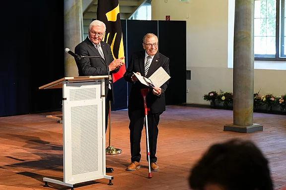 Zwei Männer stehen auf einer Bühne in einem festlichen Raum. Einer der Männer, älter und mit Brille, hält ein Dokument in der Hand und lächelt. Der andere Mann, ebenfalls älter, applaudiert. Im Hintergrund ist eine deutsche Flagge sichtbar.