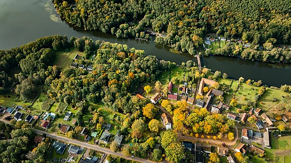 Luftaufnahme eines kleinen Dorfes, umgeben von üppigem grünem Wald und buntem Herbstlaub. Ein Fluss schlingt sich durch die Landschaft. Die Landwirtschafts- und Wohngebäude sind in harmonischer Anordnung sichtbar, die ruhige, ländliche Atmosphäre vermittelt einen Eindruck von Naturverbundenheit.