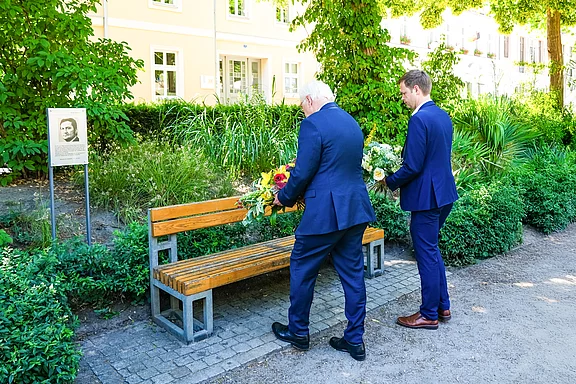 Zwei Männer in Anzügen stehen nebeneinander und legen Blumen auf eine Bank. Neben der Bank befindet sich ein Schild mit einem Foto und Informationen über eine Person. Die Umgebung ist grün mit Pflanzen und Büschen.