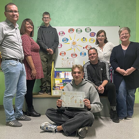 Gruppe von sieben Personen in einem Klassenraum; im Vordergrund sitzt ein junger Mann am Boden und hält eine Urkunde oder ein Arbeitsblatt hoch. Hinter ihnen steht ein Whiteboard mit farbiger Übersicht und Lernmaterialien, was auf ein Schulprojekt oder eine Präsentation hinweist.