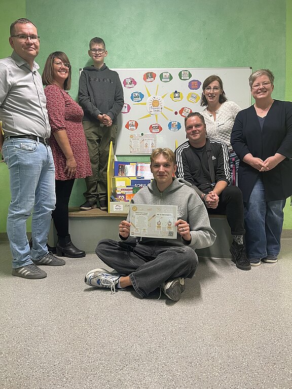 Gruppe von sieben Personen in einem Klassenraum; im Vordergrund sitzt ein junger Mann am Boden und hält eine Urkunde oder ein Arbeitsblatt hoch. Hinter ihnen steht ein Whiteboard mit farbiger Übersicht und Lernmaterialien, was auf ein Schulprojekt oder eine Präsentation hinweist.