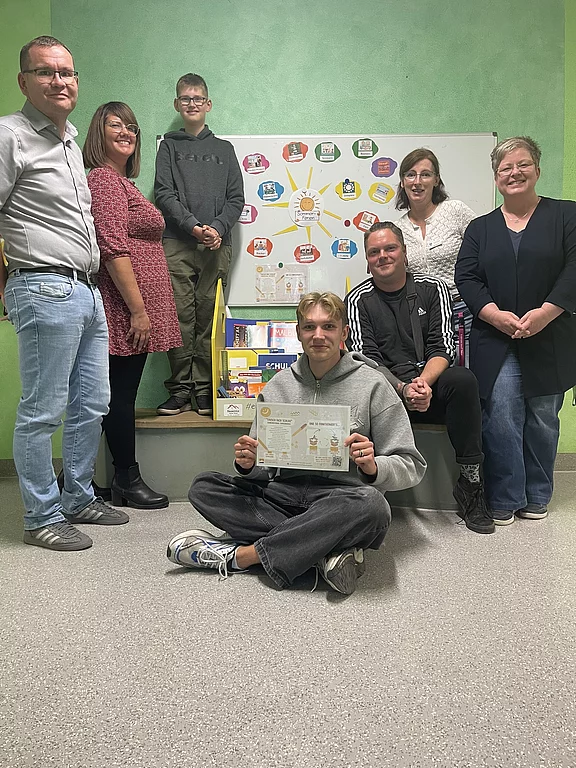 Gruppe von sieben Personen in einem Klassenraum; im Vordergrund sitzt ein junger Mann am Boden und hält eine Urkunde oder ein Arbeitsblatt hoch. Hinter ihnen steht ein Whiteboard mit farbiger Übersicht und Lernmaterialien, was auf ein Schulprojekt oder eine Präsentation hinweist.