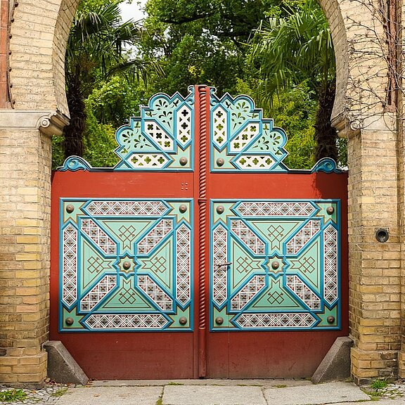 Prunkvolles, symmetrisches Tor in einem Backsteinbogen. Die zweiflügelige Holztür ist in Rot, Türkis und Weiß mit geometrischen Mustern verziert und besitzt dekorative Aufsätze. Dahinter stehen grüne Palmen und Bäume, die auf einen Garten oder Park hinweisen.