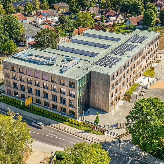 Das Bild zeigt ein modernes, mehrstöckiges Gebäude mit einer grünen Dachfläche, auf der Solarpanels installiert sind. Um das Gebäude herum befinden sich Straßen und Bäume. Es liegt in einer städtischen Umgebung mit weiteren Wohnhäusern im Hintergrund.
