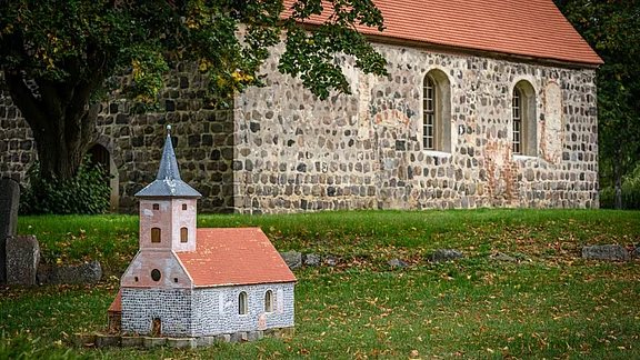 Ein kleines Modell einer Kirche mit einem grauen Steinfundament und einem roten Dach steht im Vordergrund. Im Hintergrund befindet sich eine reale Kirche aus grauem Stein mit Fenstern, umgeben von grünem Gras und Bäumen.