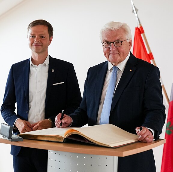 Der Bundespräsident steht an einem Tisch und schreibt in ein großes Buch. Neben ihm steht ein weiterer Mann, der lächelt. Im Hintergrund sind Fahnen sichtbar. Die Szene vermittelt einen offiziellen, feierlichen Moment.
