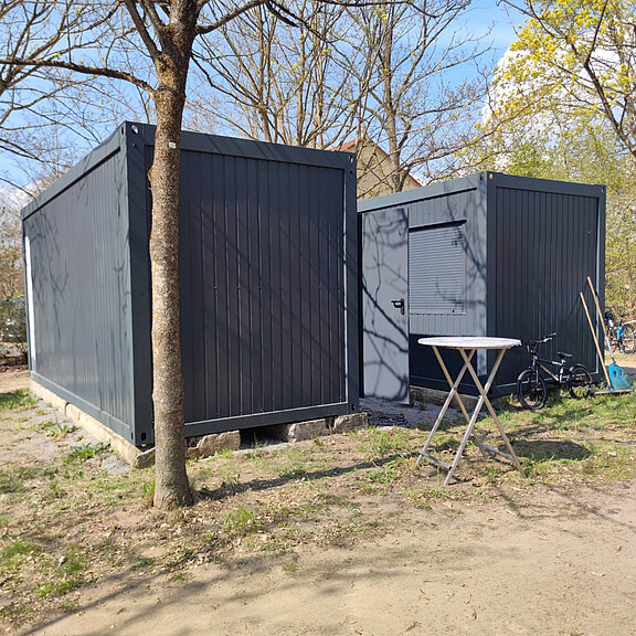 Zwei graue Container stehen neben einem Baum auf einem offenen Gelände. Der größere Container hat eine flache Dachform, während der kleinere eine Tür mit einem schmalen Fenster hat. Ein Tisch und ein Fahrrad stehen in der Nähe. Der Himmel ist klar und blau.