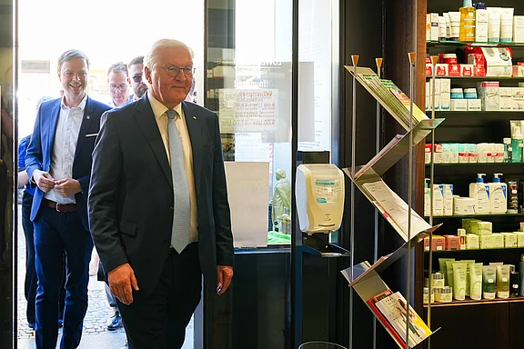Ein älterer Mann mit Brille und Anzug betritt einen Raum, gefolgt von mehreren Personen in formeller Kleidung. Im Hintergrund sind Regale mit verschiedenen Pflegeprodukten sichtbar. Der Mann wirkt freundlich und aufmerksam, während er den Raum betritt.