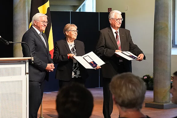 Drei Personen stehen auf einer Bühne. Sie halten Urkunden in den Händen und lächeln. Im Hintergrund ist eine Nationalflagge zu sehen. Die Szene vermittelt einen feierlichen Moment, möglicherweise eine Auszeichnung oder Ehrung.