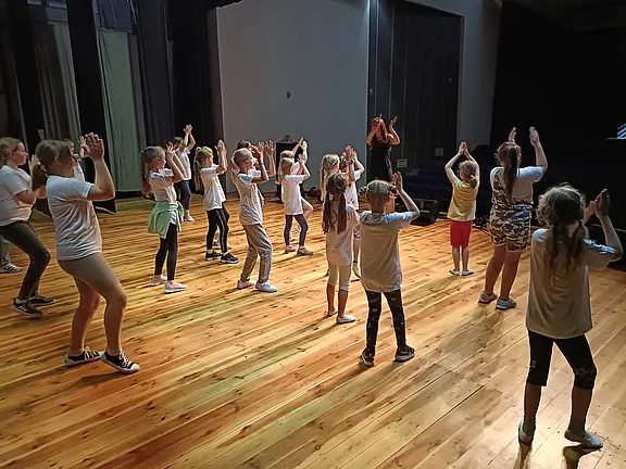 Gruppe von Kindern übt gemeinsam eine Tanzchoreografie auf einer Bühne. Sie tragen Freizeitkleidung und folgen einer Trainerin vorn, Arme hoch, in Bewegung. Holzboden, Bühnenlicht; Fokus auf gemeinsames Training und Rhythmus.
