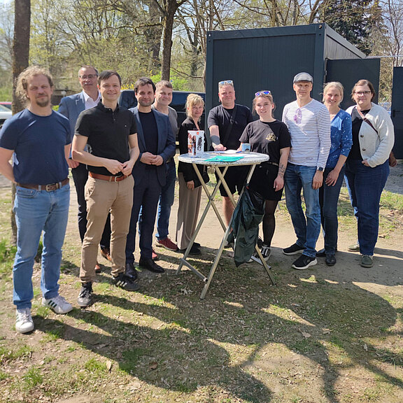 Gruppenszene im Freien mit zwölf Personen, die um einen Tisch versammelt sind. Auf dem Tisch sind Getränke und eine Torte. Die Anwesenden lächeln und stehen in lockerer Pose. Die Umgebung zeigt Bäume und einige Gebäude im Hintergrund. Es ist ein sonniger Tag.