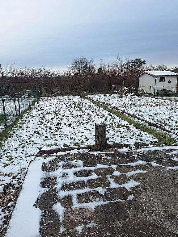 Eine Gartenlandschaft im Winter, bedeckt mit einer dünnen Schicht Schnee. Sichtbare Gartenbeete sind in parallelen Reihen angelegt. Ein Holzstumpf steht im Vordergrund, während im Hintergrund ein kleiner Schuppen und einige Bäume zu sehen sind. Der Himmel ist bewölkt.