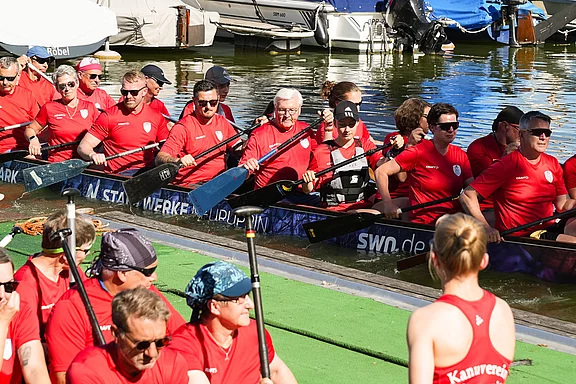 Eine Gruppe von Drachenbootfahrern in roten T-Shirts sitzt in einem Boot, bereit zum Ablegen. Im Hintergrund sind weitere Boote und Wasser zu sehen. Die Stimmung wirkt sportlich und enthusiastisch, während die Mannschaft sich auf das Rennen vorbereitet.