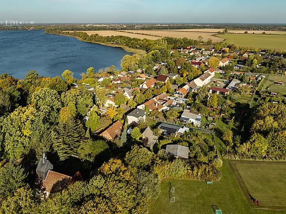 Eine Luftaufnahme eines Dorfes am Ufer eines Gewässers. Bunte Bäume umgeben die Wohnhäuser mit roten Dächern und Gärten. Im Hintergrund erstreckt sich ein weiteres Feld, während die sanften Hügel die Landschaft prägen und der Himmel klar ist.