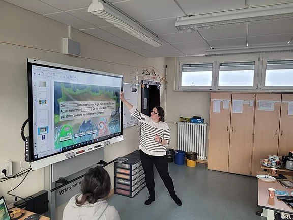 Lehrerin zeigt an einem großen interaktiven Whiteboard eine Folie über das menschliche Auge. Einige Schülerinnen und Schüler sitzen davor und folgen dem Unterricht. Digitale Präsentation in einem modernen Klassenraum.