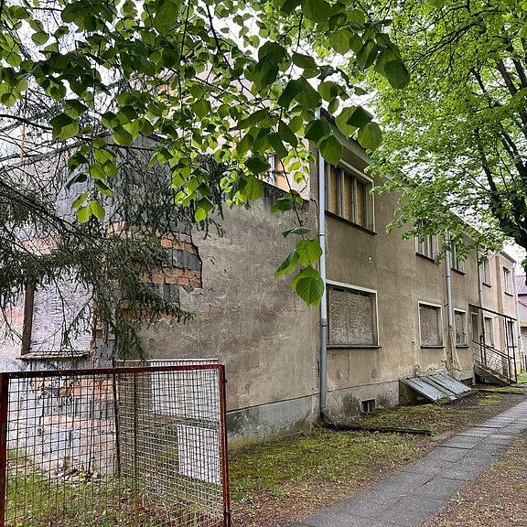 Verlassenes, verfallenes Wohngebäude an einem schmalen Gehweg, mit bröckelnder Fassade und teils vernagelten Fenstern, umgeben von Bäumen und etwas Bewuchs.