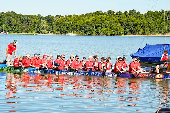 Eine Gruppe von 16 Menschen in roten T-Shirts sitzt in einem langen Rennboot auf einem Gewässer. Im Hintergrund sind Bäume und ein kleiner Bootsanleger zu sehen. Die Atmosphäre ist sonnig und zeigt eine entspannte Kajak- oder Drachenboot-Umgebung.