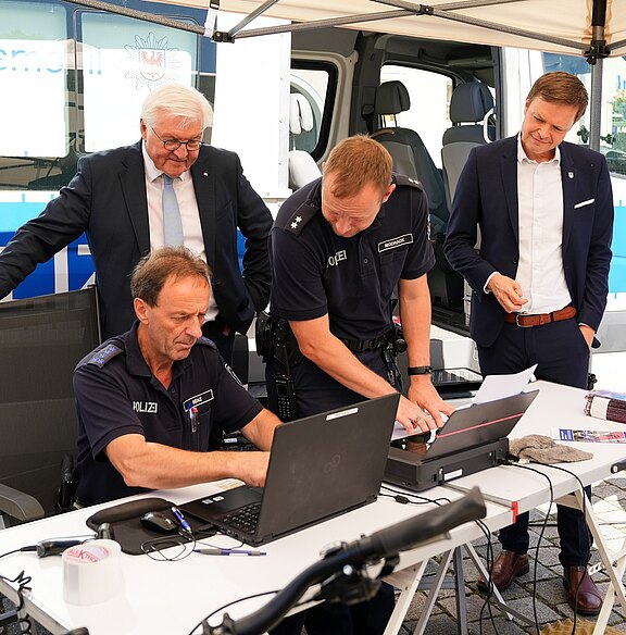 Vier Männer stehen um einen Tisch, an dem ein Polizist an einem Laptop arbeitet. Zwei der Männer tragen Polizeiuniformen, während die anderen in Anzügen gekleidet sind. Die Szene vermittelt eine Arbeitsatmosphäre im Kontext der Polizei oder öffentlichen Sicherheit.