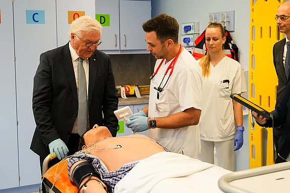 Eine Gruppe von Fachleuten in einem fiktiven Krankenhauslabor steht um ein Patientenmodell. Ein Arzt, ausgestattet mit einem Stethoskop, erklärt etwas, während ein Mann in Anzug aufmerksam zuhört. Im Hintergrund sind medizinische Geräte sichtbar.